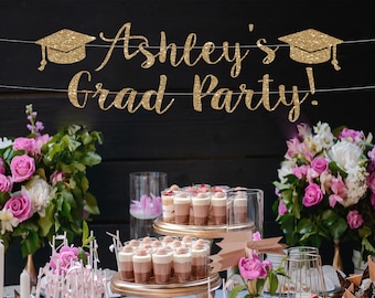 May include: Gold glitter graduation banner with the text "Ashley's Grad Party!" hanging above a dessert table with pink flowers and small desserts in clear glass cups.