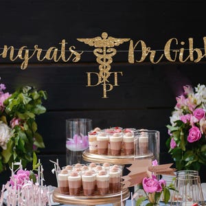 May include: Gold glitter banner with the text "Congrats Dr. Gibbs" and a medical symbol. The banner is hanging above a table with pink flowers and desserts.
