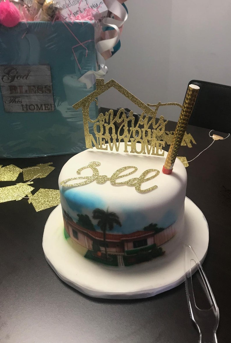 May include: A white cake with a picture of a house on it. The cake is decorated with a gold glitter topper that says "Congrats on your new home".