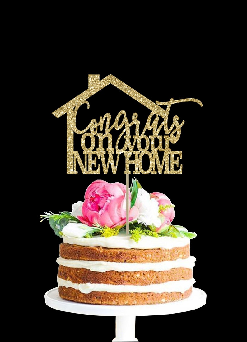 May include: Gold glitter cake topper with a house shape and the text "Congrats on your new home". The topper is on a cake with pink flowers.