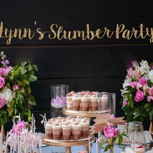 May include: A gold glitter banner with the text "Lynn's Slumber Party" suspended above a table featuring tiered dessert stands filled with layered chocolate mousse cups, surrounded by arrangements of pink and white flowers in vases.