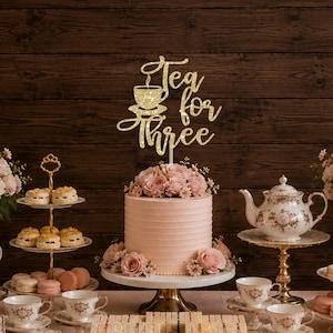 May include: A gold glitter cake topper featuring the text Tea for three with a graphic of a teacup on a saucer. The topper is positioned above a light pink frosted cake decorated with pink roses and small white flowers on a cake stand.