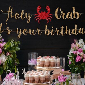 May include: A gold glitter banner with the text Holy Crab it's your birthday, featuring a red glitter crab icon in the center. The letters are in a cursive script font and are strung together on a thin white string against a dark background.