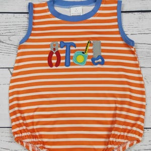 May include: A sleeveless bubble romper featuring horizontal orange and white stripes with a blue crew neckline. The centre displays an embroidered graphic of a pair of red pliers, a blue hammer, a teal tape measure, and a grey hand saw.