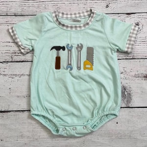 May include: A light mint green short-sleeved bubble romper featuring a grey and white gingham trim on the collar and sleeve cuffs. The centre of the chest displays an embroidered applique design of four construction tools: a hammer, two wrenches, and a saw.