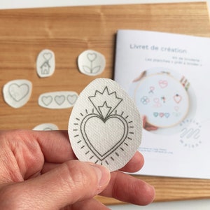 May include: A white fabric circle with a black outline of a heart with a crown and rays around it. The fabric is textured and the design is simple and elegant.