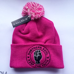 May include: A pink beanie with a white and pink pom pom on top. The beanie has a black embroidered logo that says "Northern Soul Keep The Faith" with a raised fist in the centre.
