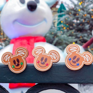 May include: Three wooden gingerbread Mickey Mouse head ornaments with white icing details and smiling faces. The ornaments are brown with green, red, and blue accents. A large snowman wearing a red scarf and hat is in the background.