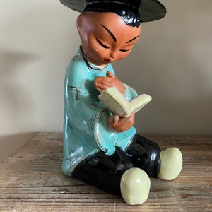 May include: A ceramic figurine of a person wearing a black hat and blue robe, sitting and reading a book. The figurine has a light brown skin tone and is painted with details.