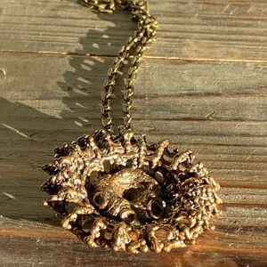 Puede incluir: Un collar dorado con un gran colgante intrincado. El colgante est&aacute; hecho de metal texturizado y tiene una forma circular. El collar est&aacute; sobre una superficie de madera.