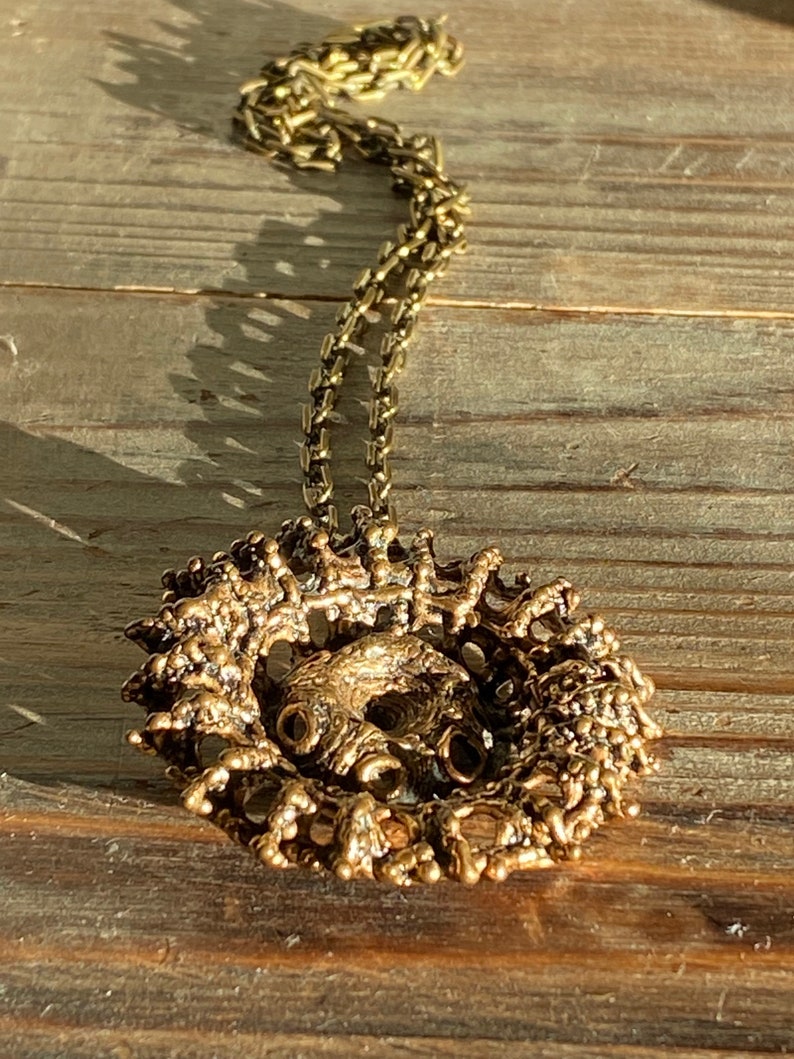 Puede incluir: Un collar dorado con un gran colgante intrincado. El colgante est&aacute; hecho de metal texturizado y tiene una forma circular. El collar est&aacute; sobre una superficie de madera.