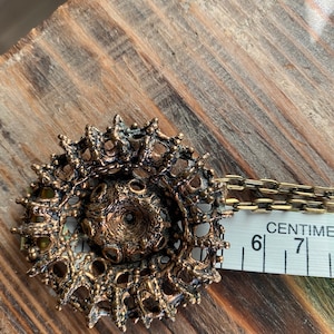 Puede incluir: Un collar con un colgante dorado de dise&ntilde;o circular intrincado. El colgante est&aacute; hecho de metal con una superficie texturizada y tiene un peque&ntilde;o agujero en el centro. El collar est&aacute; unido a una cadena.