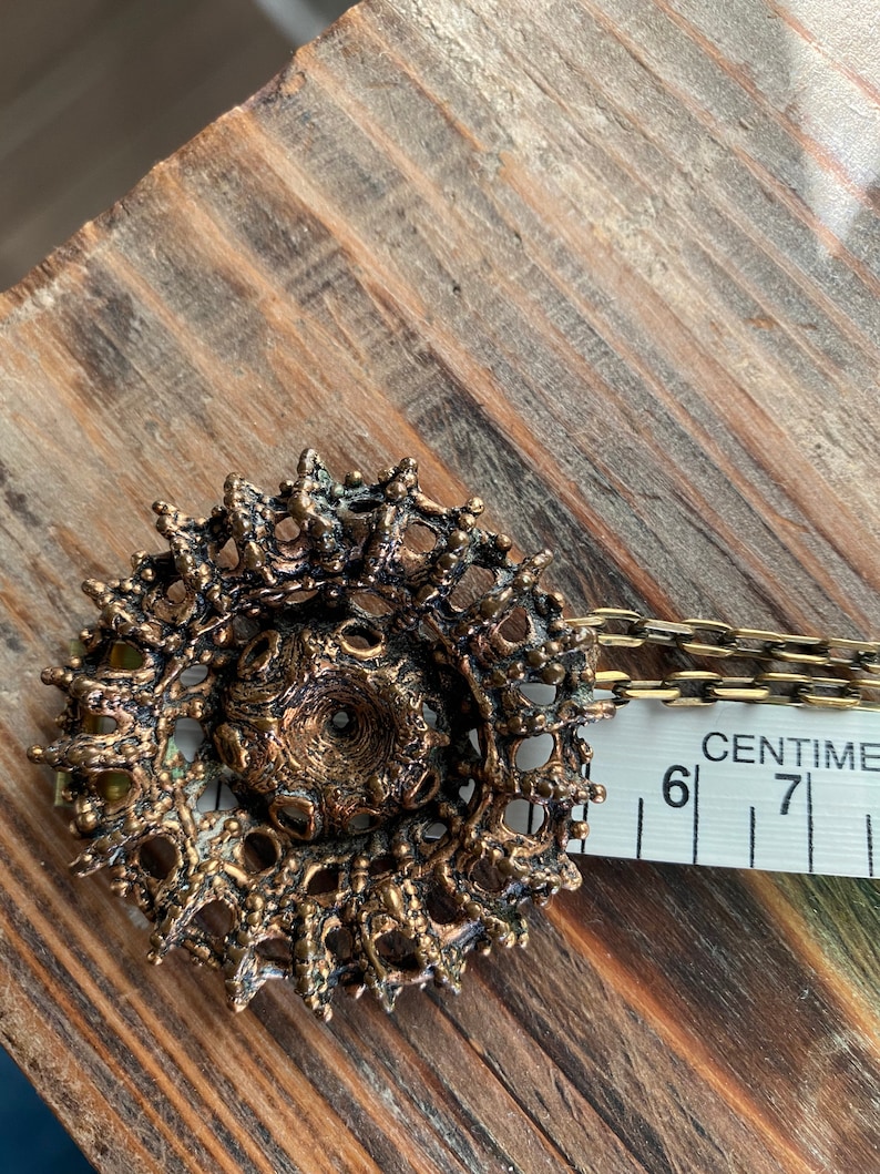 Puede incluir: Un collar con un colgante dorado de dise&ntilde;o circular intrincado. El colgante est&aacute; hecho de metal con una superficie texturizada y tiene un peque&ntilde;o agujero en el centro. El collar est&aacute; unido a una cadena.