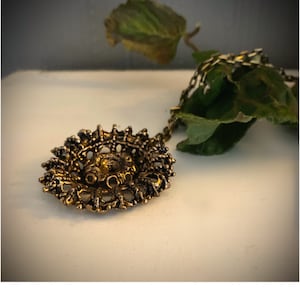 Puede incluir: Un collar con un colgante dorado de dise&ntilde;o detallado e intrincado. El colgante tiene una forma circular con un centro elevado y un delicado patr&oacute;n de filigrana.