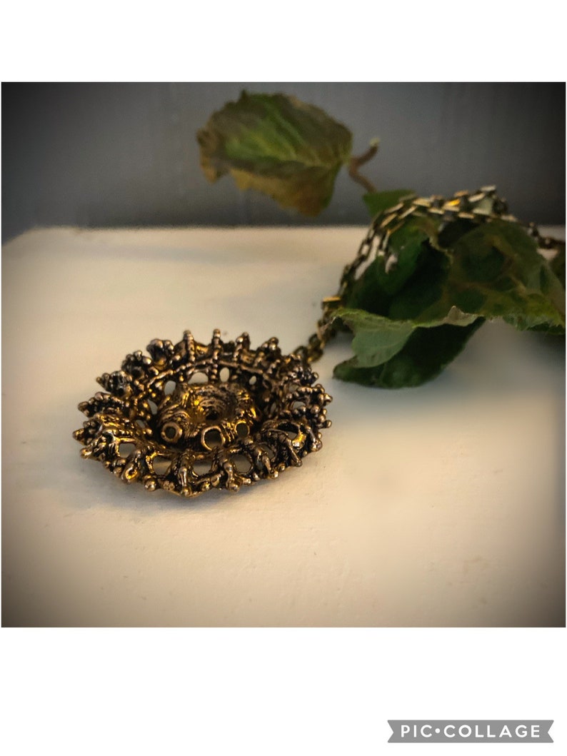 Puede incluir: Un collar con un colgante dorado de dise&ntilde;o detallado e intrincado. El colgante tiene una forma circular con un centro elevado y un delicado patr&oacute;n de filigrana.