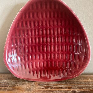 Peut inclure: Un plat en céramique triangulaire avec un motif texturé en relief. Le plat est de couleur rouge foncé.