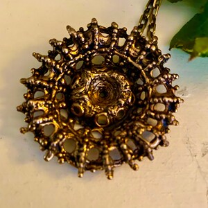 Puede incluir: Un collar con un colgante circular de color dorado, con un dise&ntilde;o intrincado y detallado. El colgante tiene un centro elevado y un delicado patr&oacute;n calado.