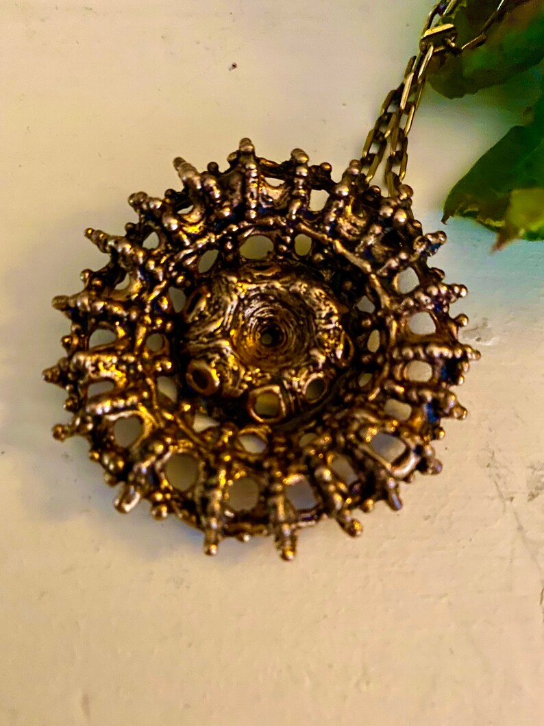 Puede incluir: Un collar con un colgante circular de color dorado, con un dise&ntilde;o intrincado y detallado. El colgante tiene un centro elevado y un delicado patr&oacute;n calado.