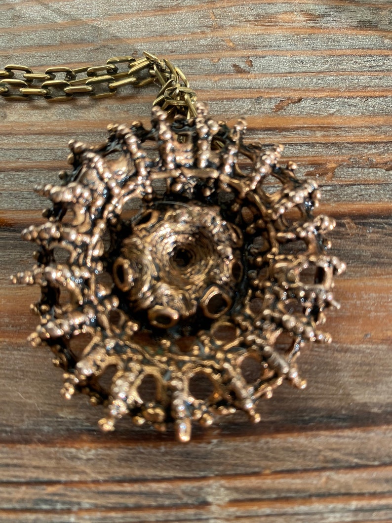 Puede incluir: Un collar con un colgante dorado de dise&ntilde;o circular intrincado. El colgante est&aacute; hecho de un metal delicado y tejido, con un aspecto texturizado y org&aacute;nico.