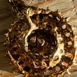 Puede incluir: Un collar con un colgante redondo de color dorado, con un dise&ntilde;o texturizado y calado. El colgante tiene una peque&ntilde;a etiqueta en la parte posterior con el texto "Studio  de  Nore".