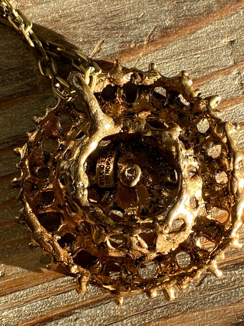 Puede incluir: Un collar con un colgante redondo de color dorado, con un dise&ntilde;o texturizado y calado. El colgante tiene una peque&ntilde;a etiqueta en la parte posterior con el texto "Studio  de  Nore".