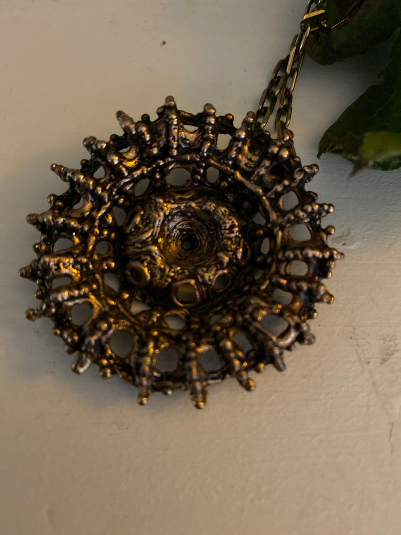 Puede incluir: Un collar con un colgante circular de color dorado, con un dise&ntilde;o intrincado y texturizado. El colgante tiene una apariencia casi org&aacute;nica con un peque&ntilde;o agujero en el centro.