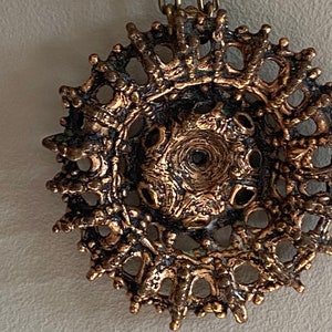 Puede incluir: Un colgante de bronce con un dise&ntilde;o circular e intrincado. El colgante tiene una superficie texturizada y un delicado patr&oacute;n calado.