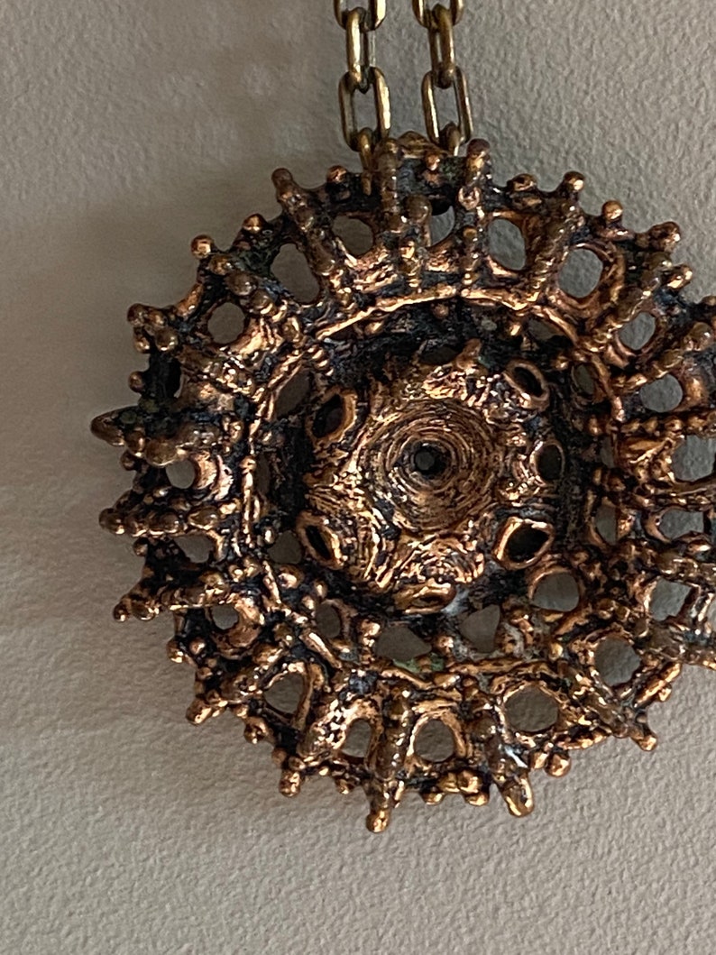 Puede incluir: Un colgante de bronce con un dise&ntilde;o circular e intrincado. El colgante tiene una superficie texturizada y un delicado patr&oacute;n calado.