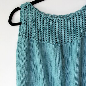 Può includere: Un top in maglia color verde acqua con uno sprone a trama aperta e strutturata. Il capo senza maniche è esposto su una gruccia nera su sfondo bianco. L'orlo inferiore è a coste. Il top è realizzato in un materiale morbido e confortevole.