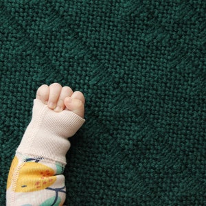 May include: A baby's hand in a pink and white long-sleeved onesie with a colorful fruit print, against a dark green knitted background.