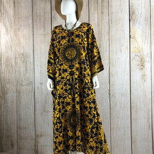 May include: A floor-length kaftan dress featuring a black base with a repeating gold ornate baroque pattern, including circular medallions. The garment has wide sleeves, a round neckline, and is displayed on a mannequin wearing a tan sun hat.
