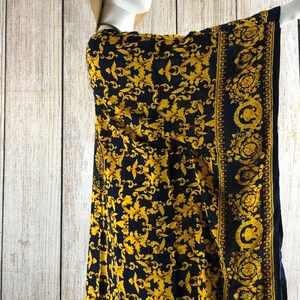 May include: A black and gold kaftan featuring an intricate repeating scrollwork pattern. The garment includes a wide border with circular motifs along the edge. The fabric is draped over a mannequin wearing a tan straw hat with a black ribbon.