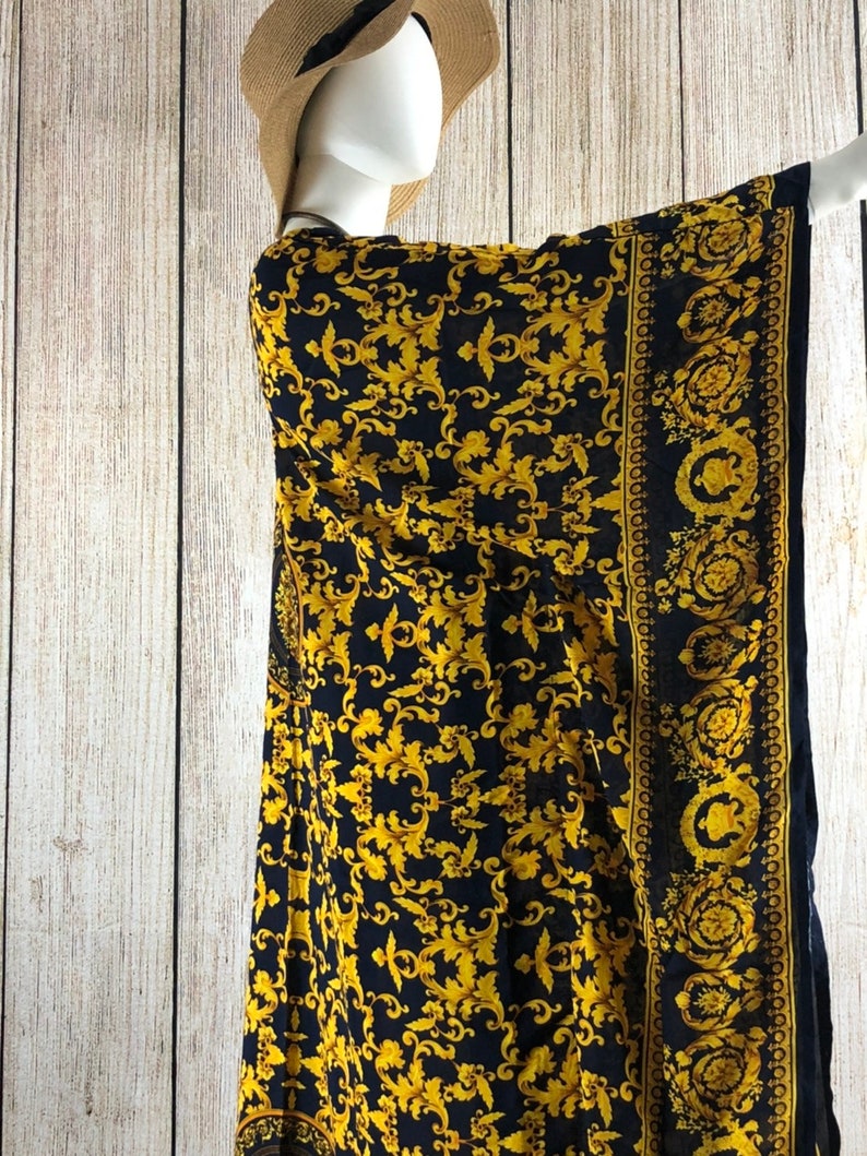 May include: A black and gold kaftan featuring an intricate repeating scrollwork pattern. The garment includes a wide border with circular motifs along the edge. The fabric is draped over a mannequin wearing a tan straw hat with a black ribbon.