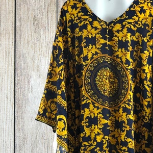 May include: A dark navy blue kaftan featuring a vibrant gold-colored baroque-inspired scrollwork pattern. The garment has a V-neckline, wide sleeves, and a prominent circular medallion design centered on the back with intricate floral detailing.
