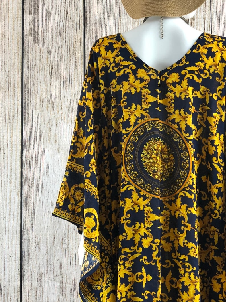 May include: A dark navy blue kaftan featuring a vibrant gold-colored baroque-inspired scrollwork pattern. The garment has a V-neckline, wide sleeves, and a prominent circular medallion design centered on the back with intricate floral detailing.
