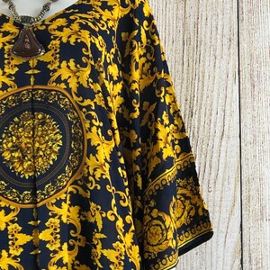 May include: A black tunic featuring a vibrant gold-colored ornate scrollwork pattern and a central circular medallion design with floral details. The garment has wide sleeves and a V-neckline, paired with a dark brown pendant necklace.