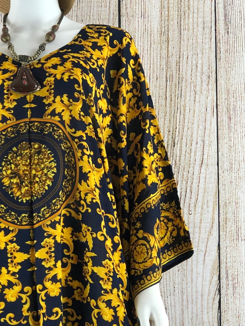 May include: A black tunic featuring a vibrant gold-colored ornate scrollwork pattern and a central circular medallion design with floral details. The garment has wide sleeves and a V-neckline, paired with a dark brown pendant necklace.