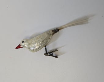 May include: A vintage-style glass bird ornament with a textured, shimmering silver finish, a small red beak, and a black eye. The bird features a long, thin, white fibre tail and is attached to a metal spring clip for secure mounting on a branch.