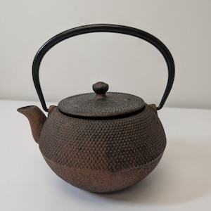 May include: A round cast iron teapot with a textured, pebbled surface in a dark brown finish. It features a matching lid with a small knob, a short spout, and a smooth, arched black metal handle that extends over the top of the teapot body.