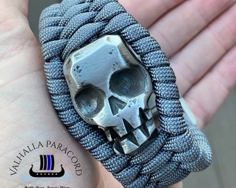 May include: A gray paracord bracelet featuring a silver skull centerpiece. The bracelet is intricately woven and has a metal skull charm. The text "VALHALLA PARACORD" and "Battle Born...Luxury Worn" are visible.