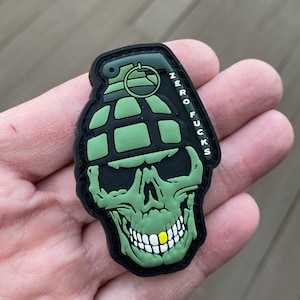 May include: A green and black skull with a green grenade on its head. The skull has a gold tooth and the text "ZERO FU**KS" is written on the side.