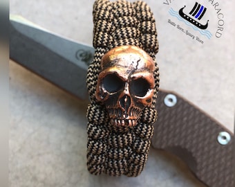 May include: A black and tan paracord bracelet with a copper skull charm. The bracelet is made with a woven pattern and has a metal clasp. The text "VALHALLA PARACORD" is visible in the background. The text "Battle Born...Luxury Worn" is also visible in the background.