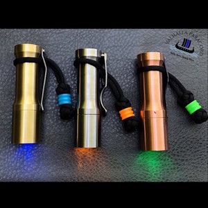May include: Three small, cylindrical flashlights with different colored lights. The flashlights are made of metal and have a black paracord lanyard attached. The flashlights are illuminated with blue, orange, and green lights.