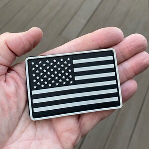 Può includere: Un patch di bandiera americana in bianco e nero con stelle e strisce bianche. Il patch è realizzato in un materiale testurizzato e ha un bordo bianco.