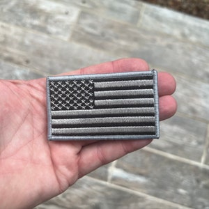 Op de afbeelding: Geborduurde patch met een ontwerp van de Amerikaanse vlag in grijstinten. De vlag heeft een gedetailleerd sterrenveld en horizontale strepen. De rechthoekige patch heeft een grijze rand, geschikt voor kleding of uitrusting.
