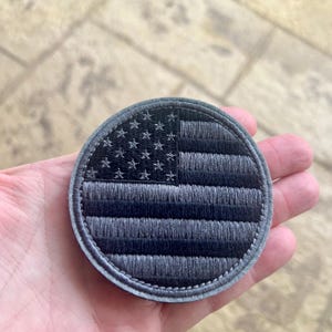 Op de afbeelding: Een ronde, geborduurde patch met een gestileerde Amerikaanse vlag in grijstinten. De patch heeft een gestikte rand en een getextureerd uiterlijk, met de sterren en strepen duidelijk gedefinieerd.