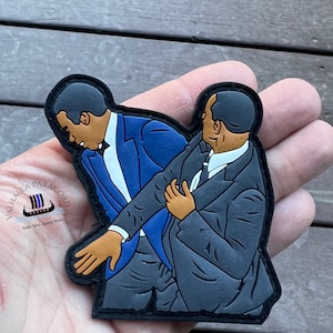 Puede incluir: Un parche bordado negro y azul con dos hombres en traje. El hombre de la izquierda lleva un traje azul y el hombre de la derecha lleva un traje negro. El parche tiene el texto "Valhalla Paracord" y "Battle Born, Always Worn" en blanco.