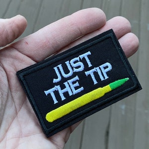 以下が含まれることがあります： 黒い長方形のパッチ。白い文字で「JUST THE TIP」と刺繍されています。黄緑色の弾丸のデザインがテキストの下に刺繍されています。パッチには黒い縁取りがあります。