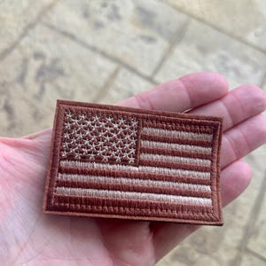 Op de afbeelding: Een bruin geborduurd patch met een wit Amerikaans vlagontwerp. De vlag heeft 13 strepen en 50 sterren.