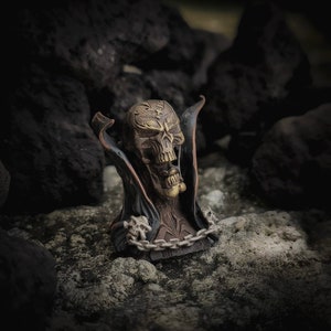 May include: A dark bronze figurine of a skull with a cape and chain. The skull has a detailed design and is set against a dark background.